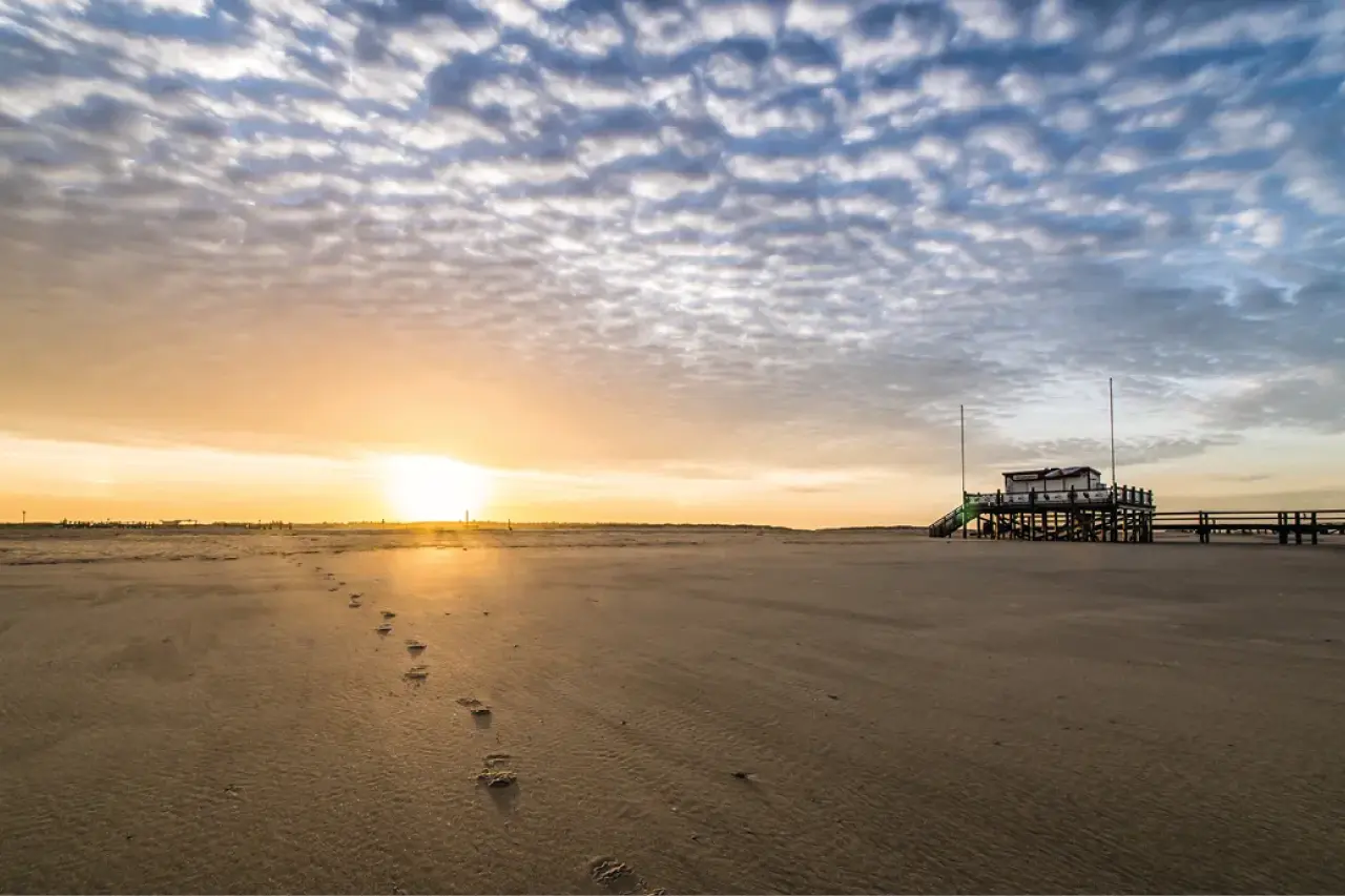 Weitläufiger Nordseestrand bei Sonnenaufgang mit Fußspuren im Sand, Pfahlbau am Horizont und strukturierter Wolkendecke, ruhige Küstenlandschaft und maritimes Urlaubsgefühl